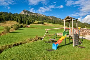 Spielplatz mit Rutsche, Schaukel und Klettergerüst vor grüner Wiese und bewaldetem Berg