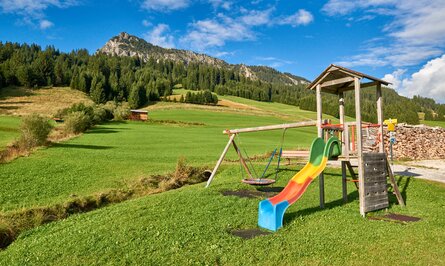 Spielplatz mit Rutsche, Schaukel und Klettergerüst vor grüner Wiese und bewaldetem Berg