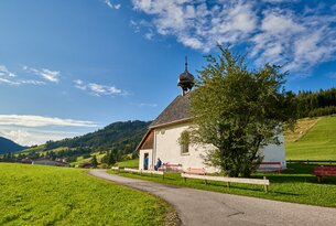 Kleine weiße Kapelle auf grüner Wiese vor Berglandschaft unter blauem Himmel