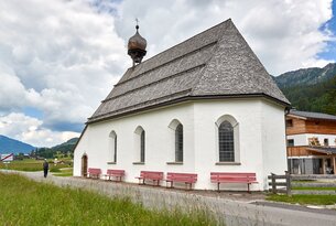 Weiße Kapelle mit rotem Bänken vor grauem Dach und bewölktem Himmel