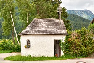 Weiße Kapelle mit Holzschindeldach vor Bäumen und Bergen
