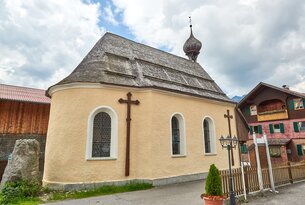 Gelbe Kapelle mit Holzkreuzen und grauem Schindeldach vor bewölktem Himmel