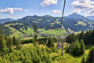 Sessellift mit zwei Personen vor Berglandschaft mit Wiesen und Wald