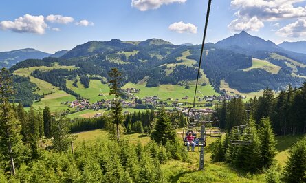 Sessellift mit zwei Personen vor Berglandschaft mit Wiesen und Wald