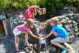 Drei Kinder spielen an einem kleinen Wasserfall mit Eimern und Bechern