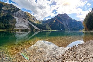 Bergsee mit Spiegelung, bewaldeten Hängen und hellem Ufer im Vordergrund
