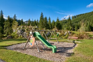 Spielplatz mit Rutsche, Schaukeln und Bank vor Wald und blauem Himmel