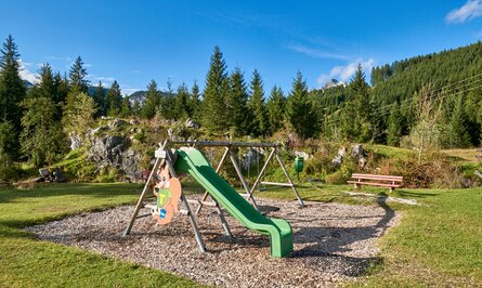 Spielplatz mit Rutsche, Schaukeln und Bank vor Wald und blauem Himmel