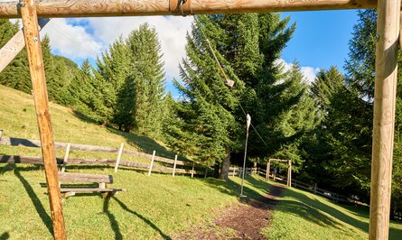 Zipline-Parcours in grüner Landschaft mit Bäumen, Zaun und Bank unter blauem Himmel