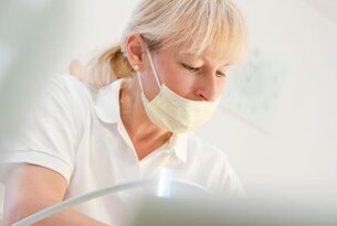 Blonde Frau mit Maske und weißem Oberteil beugt sich über ein Gerät