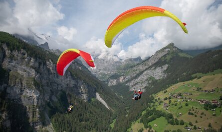 Zwei Gleitschirmflieger vor Berglandschaft mit grünen Hängen und schneebedeckten Gipfeln unter bewölktem Himmel