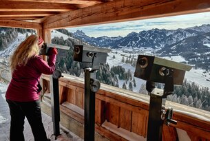 Frau blickt durch Fernrohr auf verschneite Berglandschaft