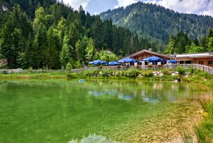 Bergsee mit Hütte, blauen Sonnenschirmen und bewaldeten Bergen im Hintergrund