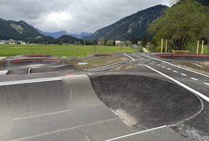 Pumptrack-Anlage vor Bergkulisse mit grüner Wiese und bewölktem Himmel