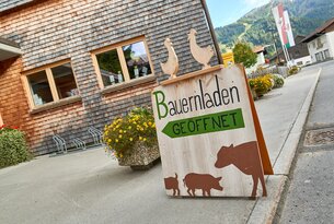 Bauernladen-Schild mit Holzfiguren, Kuh, Schwein und Ferkel vor einem Holzhaus