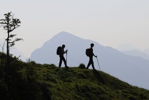 Zwei Wanderer mit Rucksäcken und Stöcken auf einem Hügel vor Bergkulisse