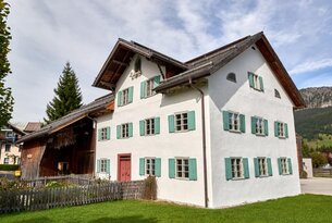 Weißes Bauernhaus mit grünen Fensterläden und roter Tür unter blauem Himmel