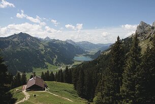 Berglandschaft mit See, bewaldeten Hängen und Hütte im Vordergrund