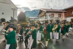 Musikkapelle in Tracht marschiert durch Dorfstraße vor Häusern und Bergen