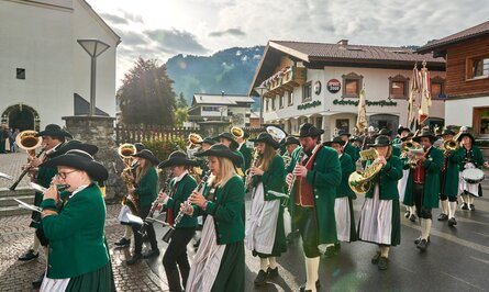Musikkapelle in Tracht marschiert durch Dorfstraße vor Häusern und Bergen