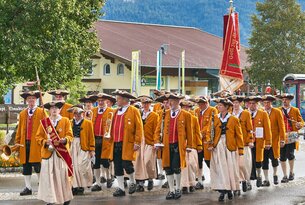 Trachtengruppe marschiert mit Instrumenten und Fahne durch eine Ortschaft
