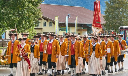 Trachtengruppe marschiert mit Instrumenten und Fahne durch eine Ortschaft