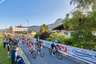 Radrennen auf Asphaltstraße vor Häusern und Bergkulisse