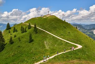 Grüner Hügel mit Wanderweg zum Gipfelkreuz unter blauem Himmel mit Wolken