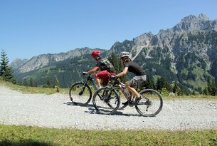 Zwei Mountainbiker fahren auf Schotterweg vor Bergpanorama