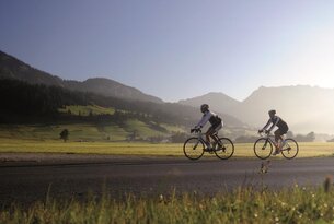 Zwei Radfahrer auf Asphaltstraße vor grüner Landschaft mit Bergen im Hintergrund