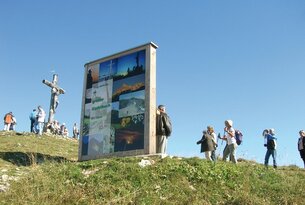 Infotafel auf Hügel mit Kreuz und Wanderern vor blauem Himmel