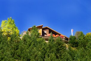 Holzhaus mit Terrasse auf einem Hügel, Bäume im Vordergrund, blauer Himmel