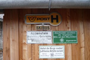 Schilder an Holzwand: Post, Skibus, Aggenstein, Alpenverein, Berge sauber halten