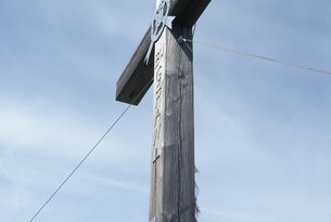 Hölzernes Gipfelkreuz mit Metallornament vor blauem Himmel