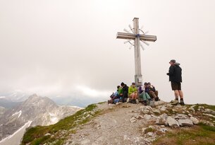 Gruppe sitzt am Gipfelkreuz auf einem Berg bei bewölktem Himmel
