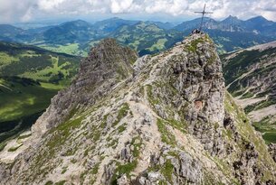 Bergpfad führt zu Gipfelkreuz auf felsigem Berggipfel mit grüner Landschaft im Hintergrund