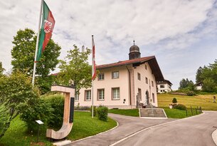 Helles Gebäude mit Fahnen und Holzskulptur am Wegesrand
