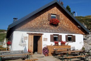 Hütte mit Holzschindeln, weißen Wänden, Blumenkästen und Holzbänken vor dem Eingang
