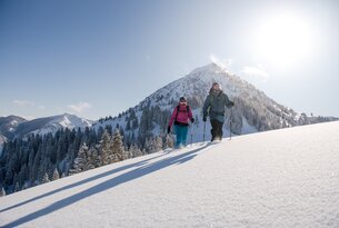 Zwei Personen wandern mit Schneeschuhen auf einem schneebedeckten Hügel vor einem Berg