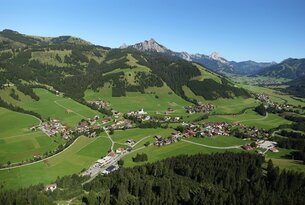 Luftaufnahme eines grünen Tals mit Dorf, umgeben von bewaldeten Hügeln und Bergen unter blauem Himmel