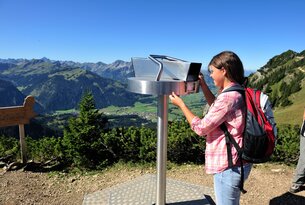 Frau mit Rucksack blickt durch ein Fernrohr auf eine Berglandschaft