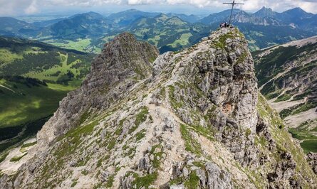 Bergspitze mit Gipfelkreuz und Wanderern, steiniger Pfad, grüne Landschaft im Hintergrund