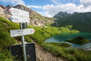 Wegweiser am Bergsee mit grünen Wiesen und Bergen im Hintergrund