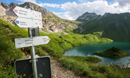 Wegweiser am Bergsee mit grünen Wiesen und Bergen im Hintergrund
