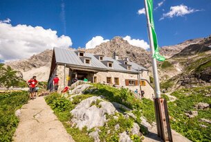 Berghütte aus Stein mit grauem Dach vor Bergpanorama und blauem Himmel