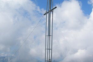 Metallkreuz auf Berggipfel, Menschen sitzen im Hintergrund, bewölkter Himmel