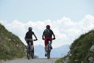 Two mountain bikers ride uphill on a gravel road against a mountain backdrop
