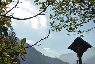 Berglandschaft mit Kreuzwegstation unter Bäumen bei sonnigem Himmel