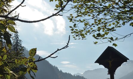 Berglandschaft mit Kreuzwegstation unter Bäumen bei sonnigem Himmel
