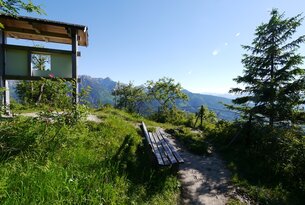 Berglandschaft mit Hütte, Weg und Bank im Vordergrund bei sonnigem Wetter